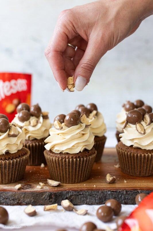 A hand crumbling Maltesers onto cupcakes