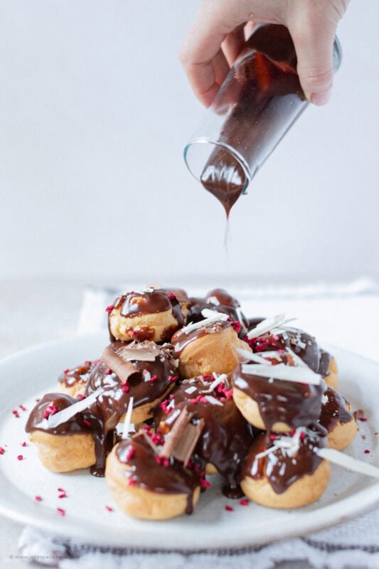 A hand pouring chocolate sauce over a plate of profiteroles