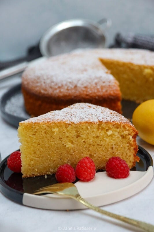 A slice of Lemon Olive Oil Cake on a plate with a fork and surrounding raspberries.